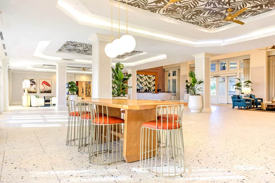 Modern hotel lobby with wooden bar, white pendant lights, potted plants, and patterned ceiling details