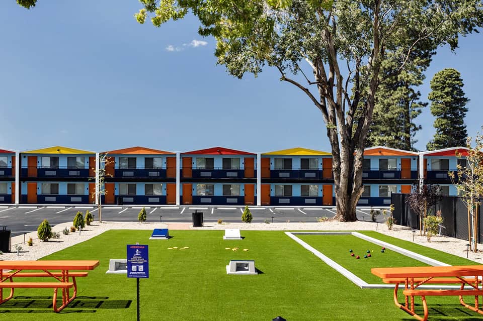Modern orange and blue residential buildings with pitched roofs overlook a manicured lawn with picnic tables and large trees