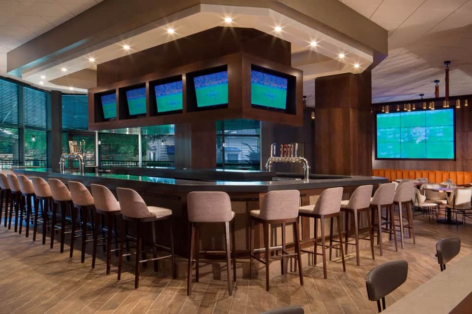 Modern sports bar with multiple screens, dark wood bar counter, and upholstered seating with ocean views