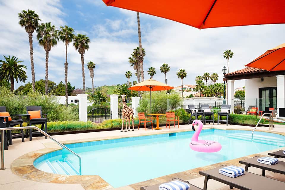 Resort pool deck with palm trees, orange umbrellas, pink flamingo float, and landscaped grounds
