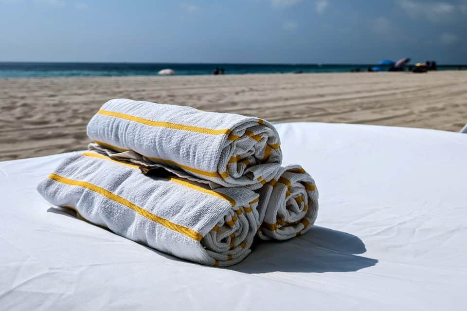 Rolled white towels with yellow stripes stacked on sandy beach with ocean and sky in background