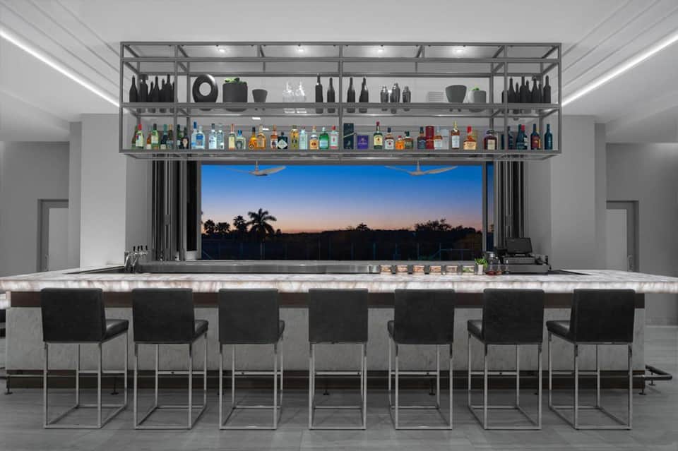 Upscale home bar with metal shelving, marble countertop, black bar chairs, and panoramic window view at dusk
