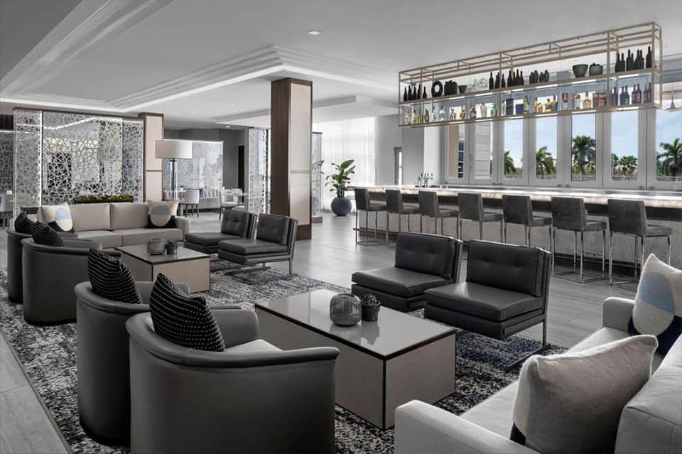 Luxury lounge with modern furniture, bar counter, and floor-to-ceiling windows overlooking palm trees