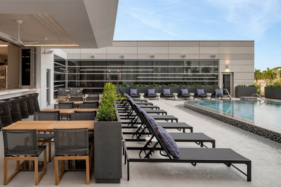 Modern rooftop pool deck with loungers, glass building facade, and palm trees under blue sky