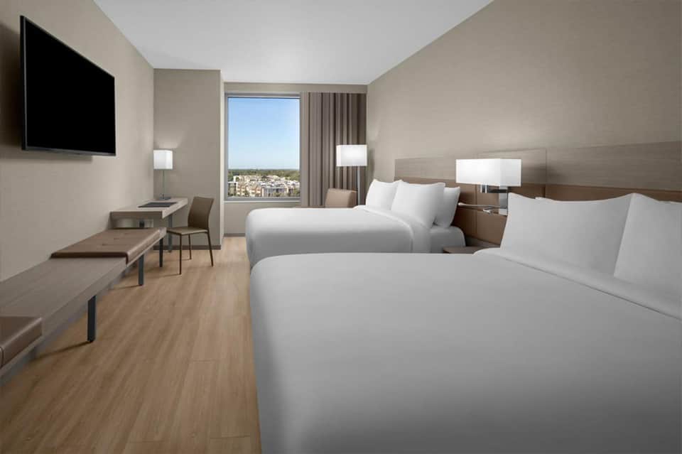 Modern hotel room with two queen beds, flat-screen TV, desk, and floor-to-ceiling window with city view