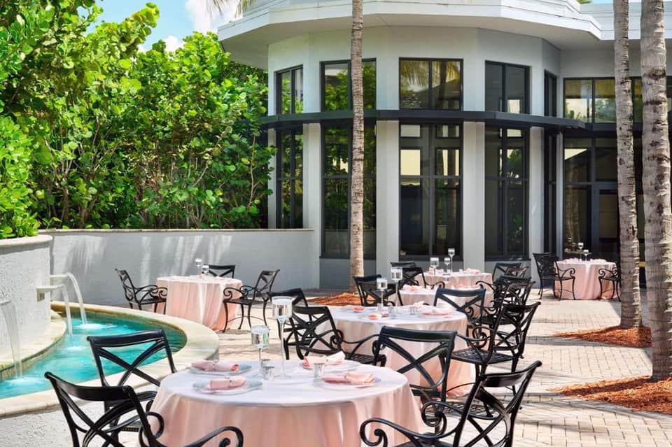 Elegant outdoor patio with round tables, pink tablecloths, black chairs, pool, and modern white building with palm trees