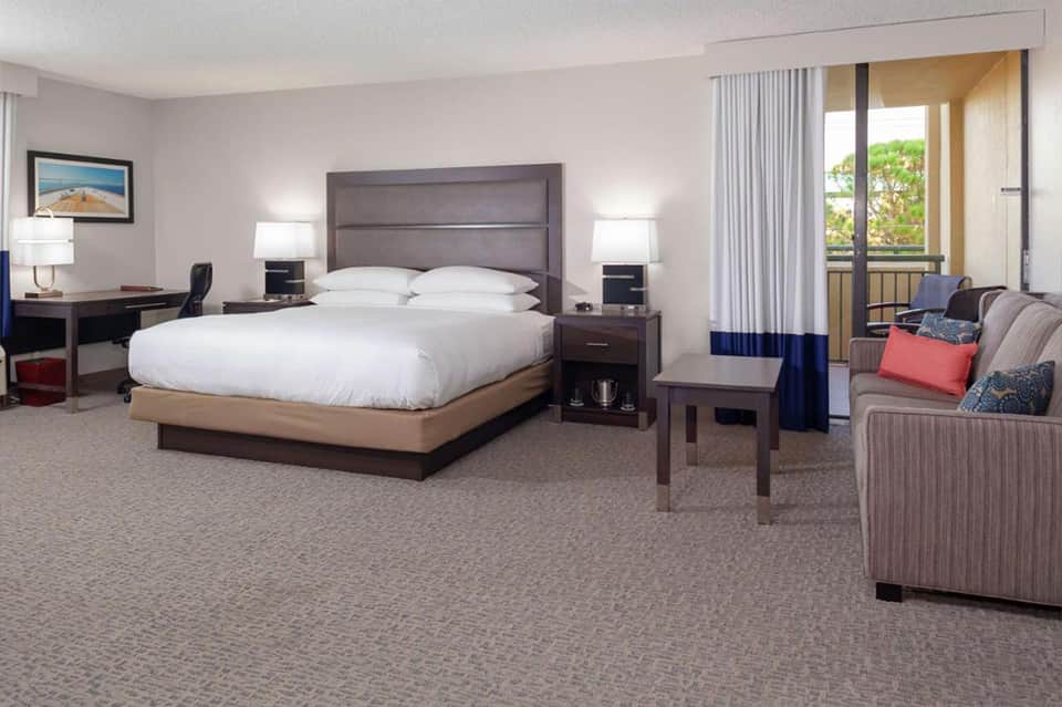 Spacious hotel suite with king bed, work desk, seating area, and balcony overlooking greenery
