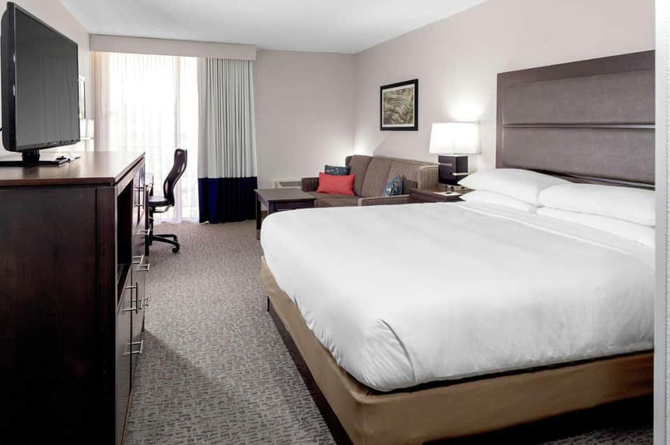 Hotel room with queen bed, gray headboard, desk, sofa, and balcony with city views
