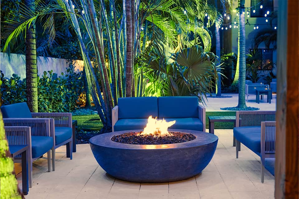 Outdoor fire pit seating area with blue chairs, palm trees, and tropical landscape lighting