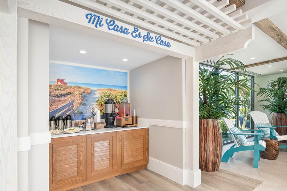 Coffee bar area with 'Mi Casa Es Su Casa' sign, wooden cabinetry, coffee maker, and view to patio with blue Adirondack chairs
