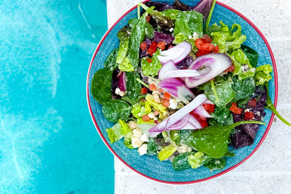 Colorful salad with mixed greens, red onions, and fresh vegetables on blue plate by pool