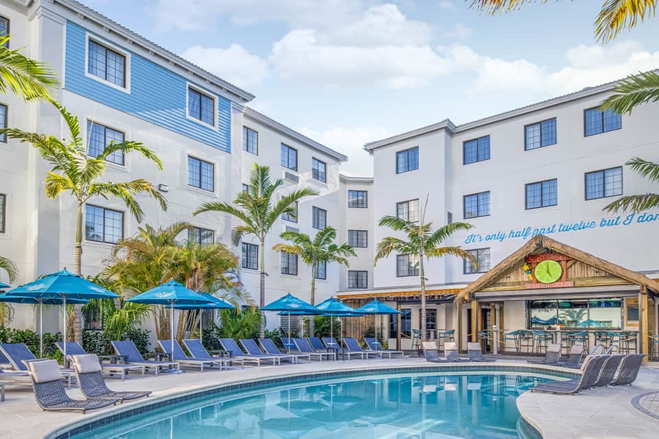 Resort pool with blue umbrellas and loungers, palm trees, hotel buildings with white walls