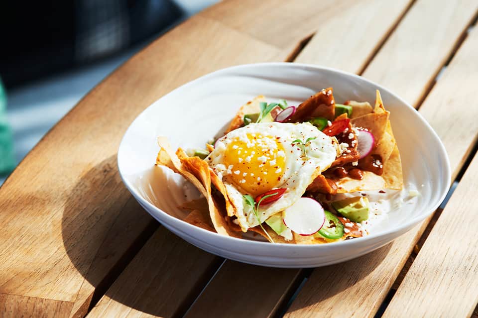 Plated dish with fried tortilla chips, fried egg, avocado, radish, and queso fresco on wooden table