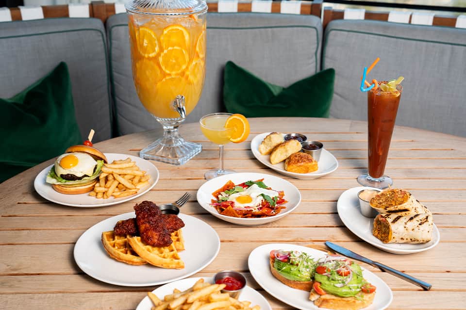 Casual brunch spread featuring waffles with bacon, egg sandwich, avocado toast, and fresh citrus beverages