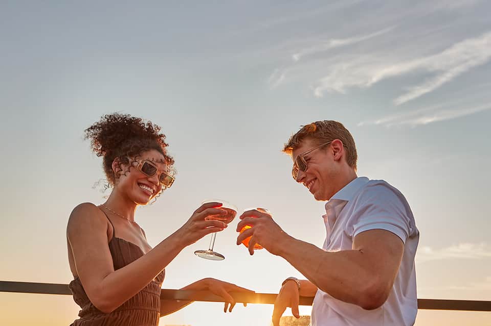 Couple toasting with drinks at sunset on a balcony overlooking the ocean