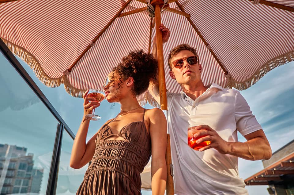 Couple enjoying cocktails under a striped umbrella on a rooftop terrace with city skyline