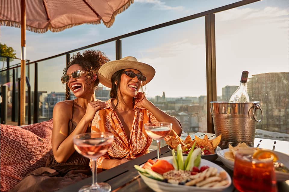 Two women enjoying cocktails on a rooftop terrace with city skyline views, appetizers, and wine bucket