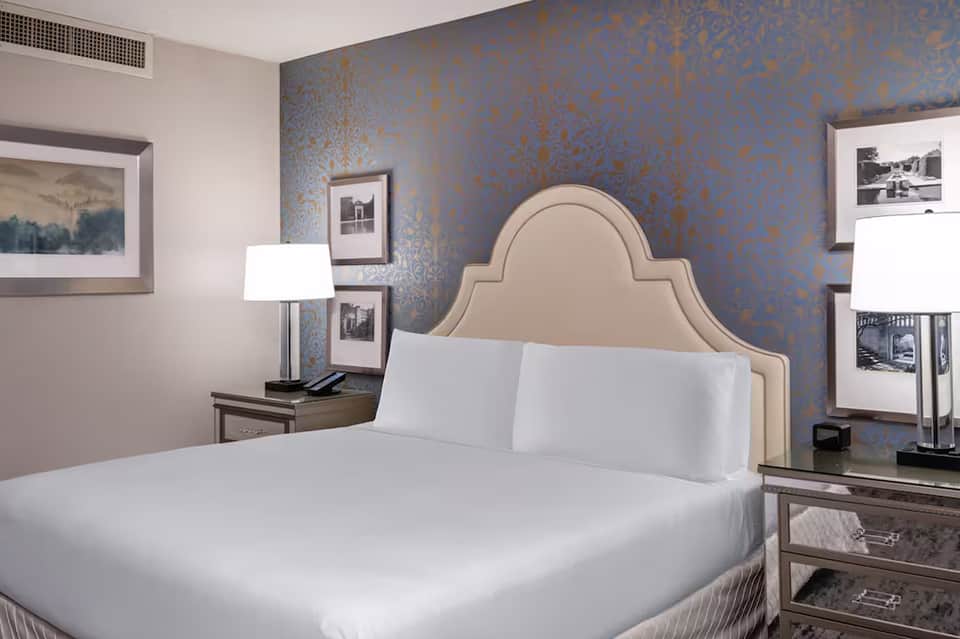 Elegant hotel bedroom with beige upholstered headboard, white bedding, blue damask wallpaper, and modern furnishings