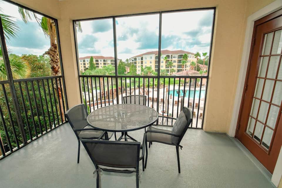 Balcony overlooking resort pool and grounds with round patio table and four chairs in afternoon sunlight.