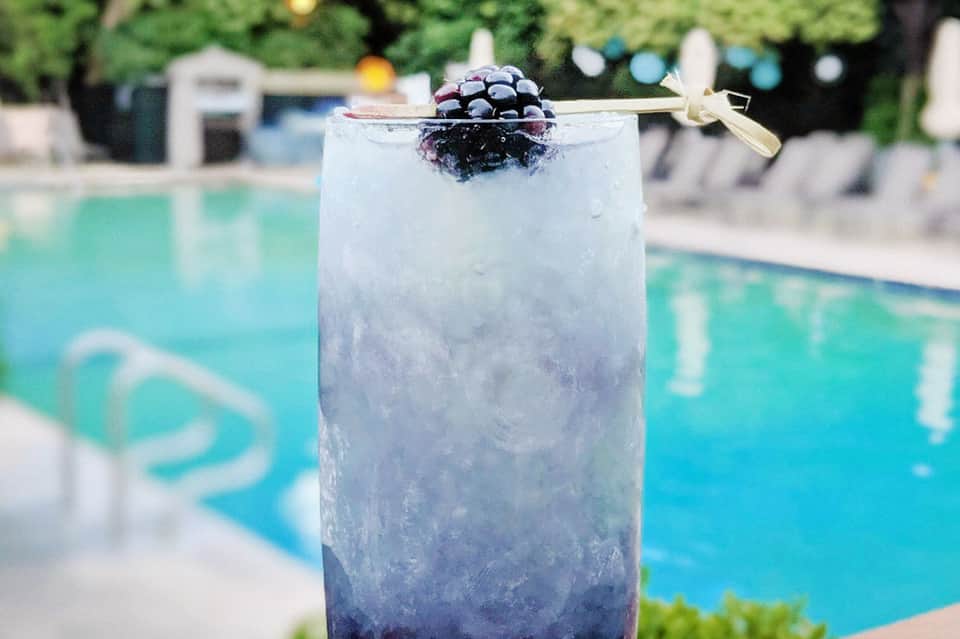 Blackberry cocktail with ice garnished with a gold bow tie, poolside with turquoise water in background