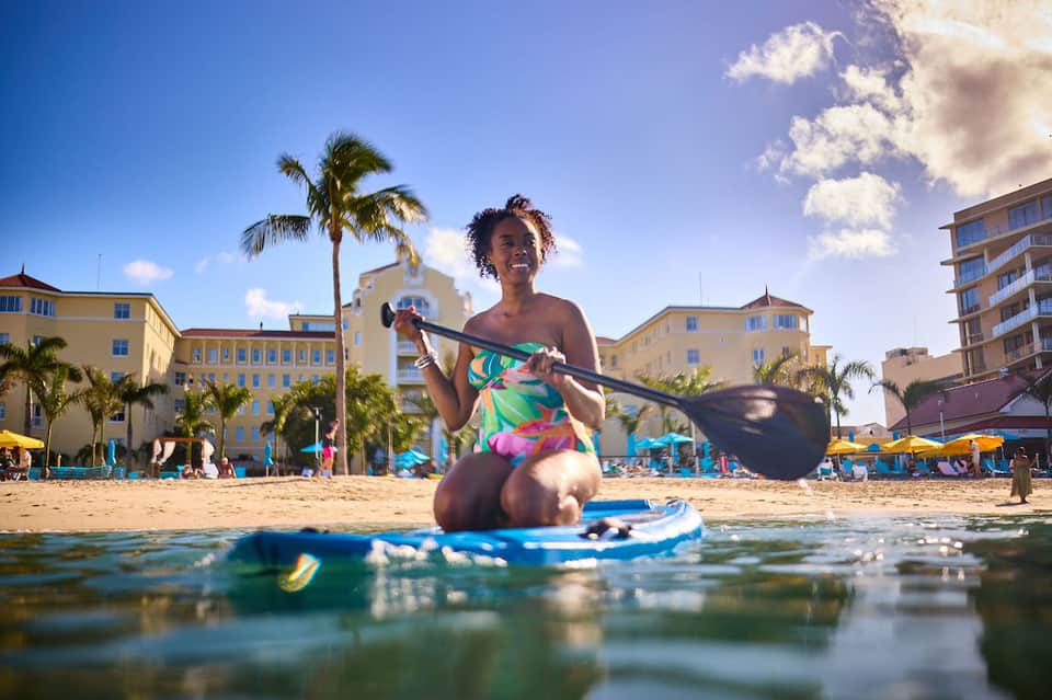 Woman on paddleboard in shallow water near beach with colorful buildings and palm trees in background