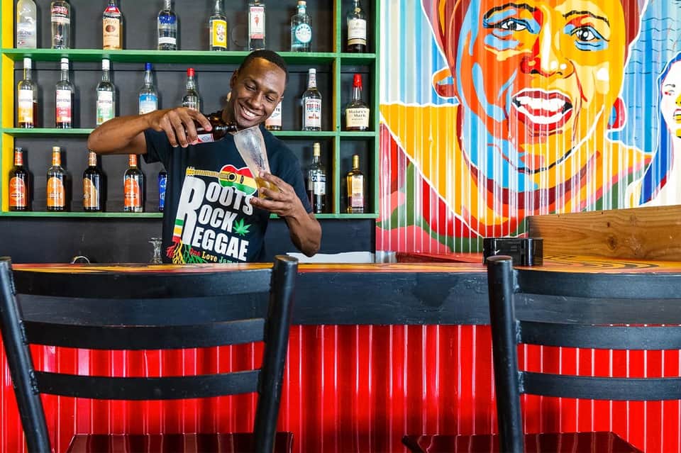 Bartender smiling at colorful bar with spirit bottles and vibrant portrait artwork