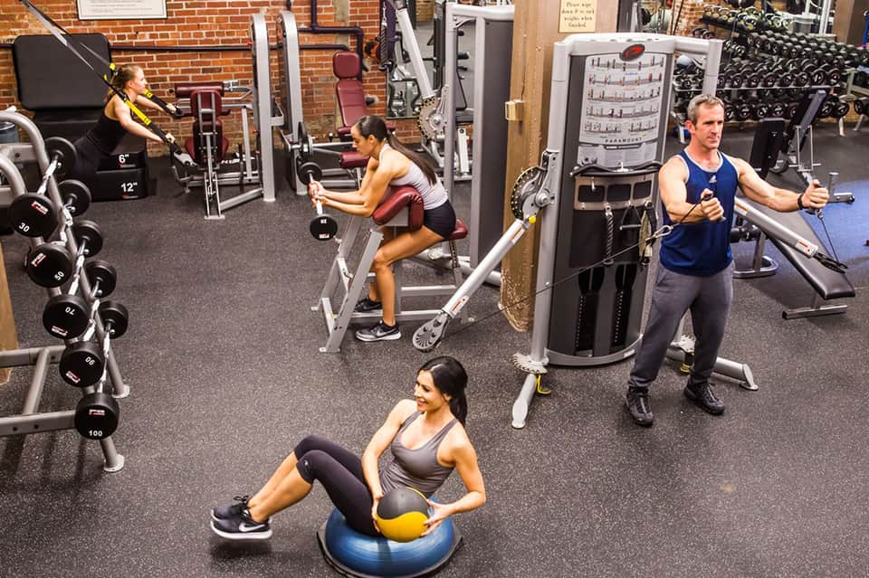 Indoor gym with fitness equipment, dumbbells, machines, and people exercising in brick-walled facility