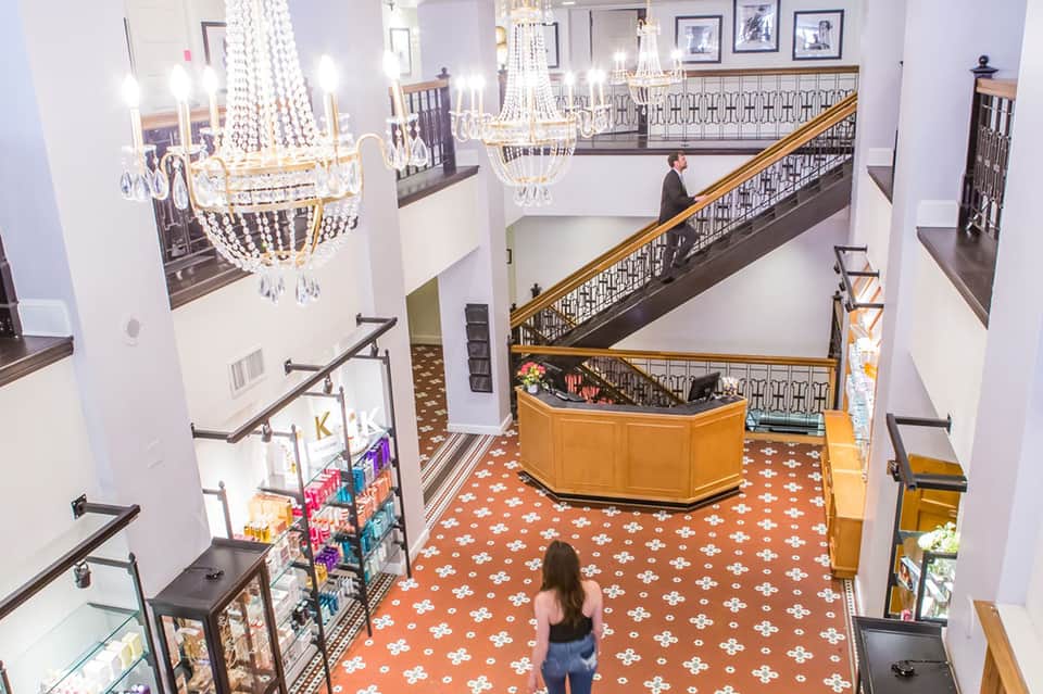 Grand hotel lobby with crystal chandeliers, multi-level wooden railings, orange patterned floor, and guest on staircase