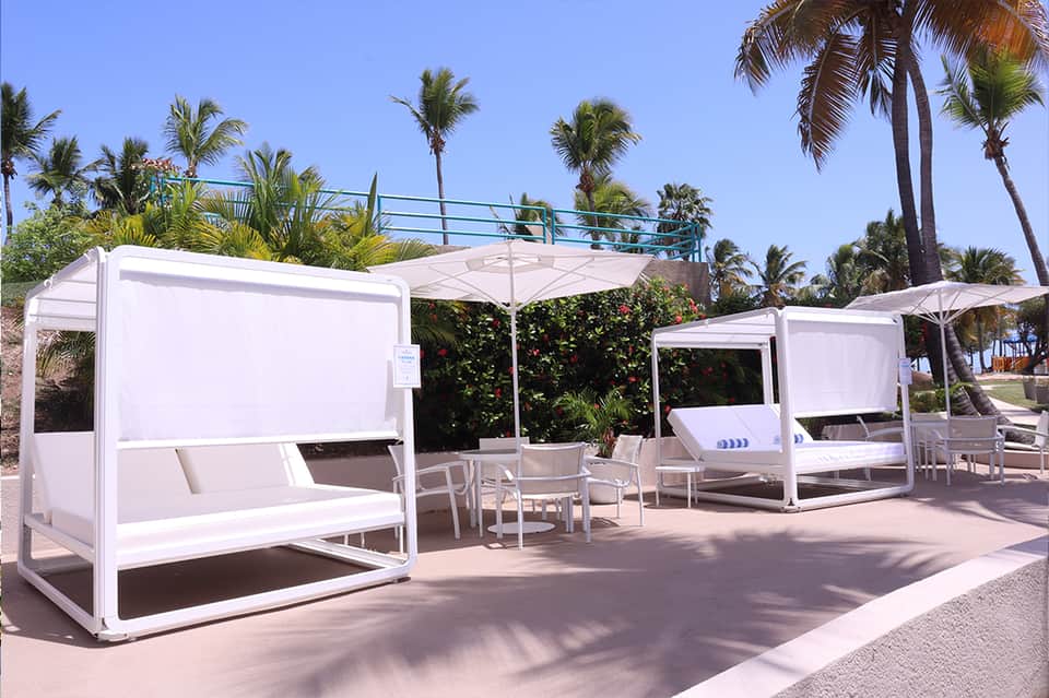 Beachside lounge area with white cabanas, umbrellas, and palm trees under clear sky