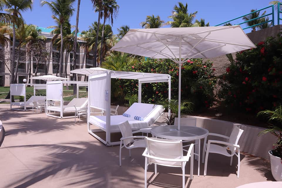 Resort pool deck with white lounge chairs, large umbrella, and tropical landscaping with palm trees