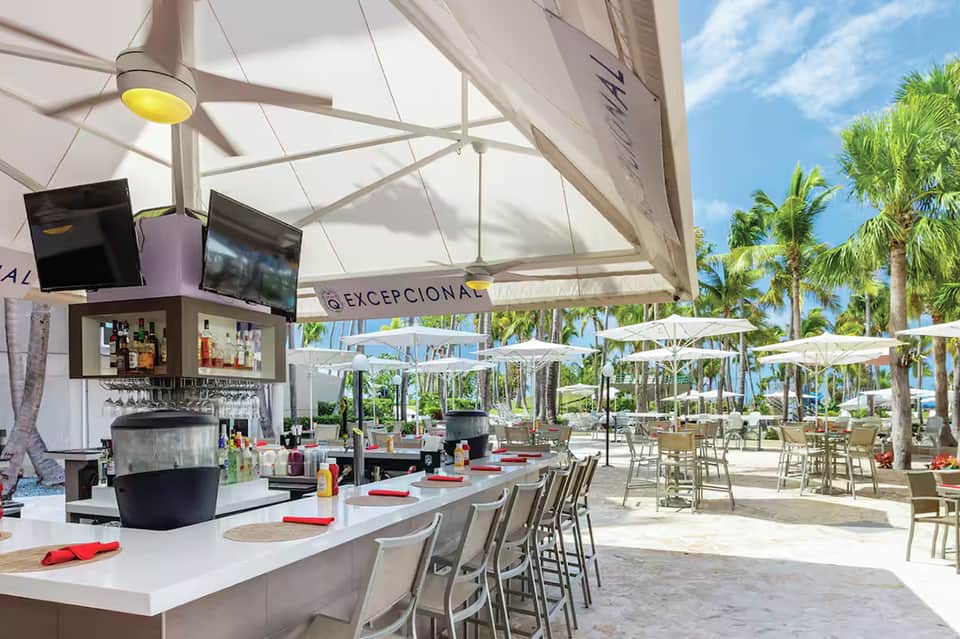 Beachfront restaurant with modern bar, white umbrellas, and palm trees under retractable shade canopy