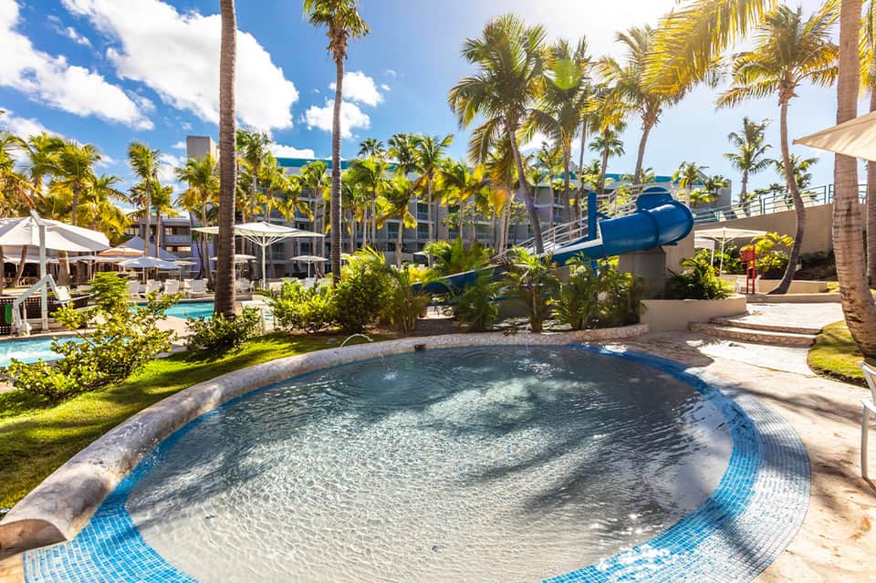 Resort pool area with palm trees, water slide, lounge chairs, and ocean views in tropical beachfront setting