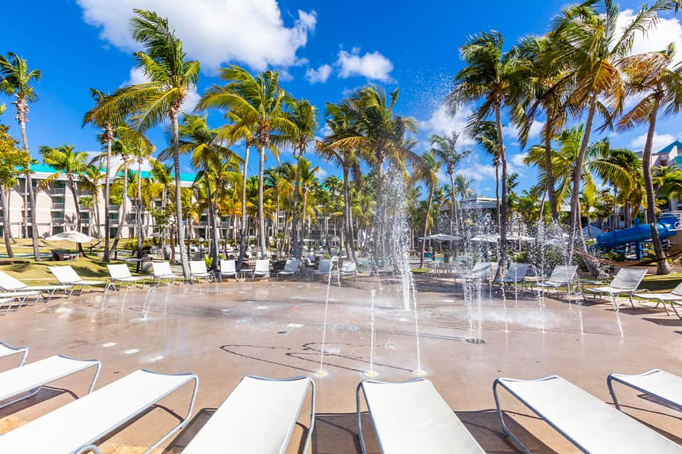 Sandy beach area with white lounge chairs, palm trees, and decorative water fountains
