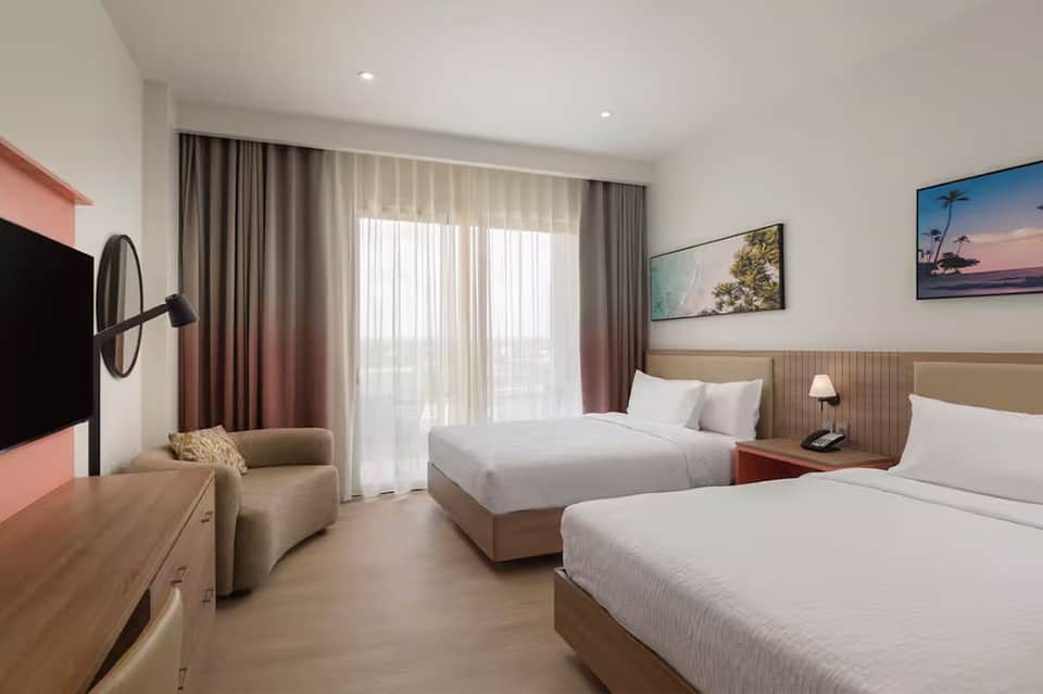 Modern hotel room with two beds, beige headboard, coral accents, and balcony access with city views