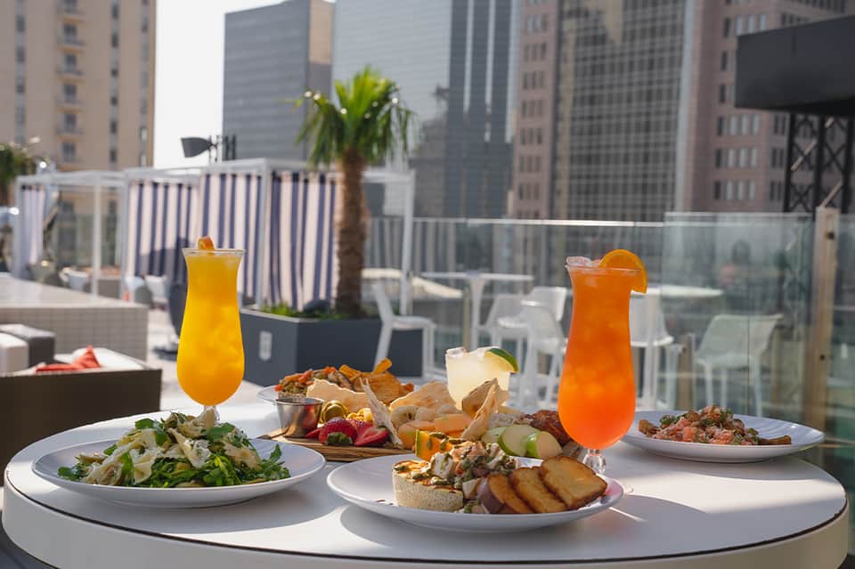 Rooftop dining table with colorful cocktails, salads, and appetizers overlooking city skyline