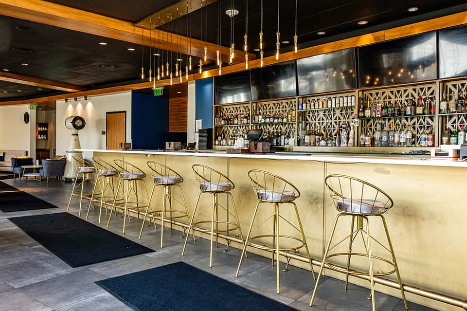 Upscale bar lounge with brass stools, geometric patterned backlit shelving, and dark ceiling with pendant lights