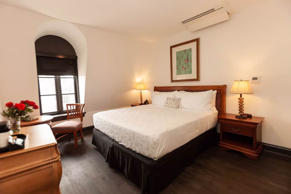 Elegant hotel bedroom with queen bed, wooden furniture, arched window with city view, and warm lighting