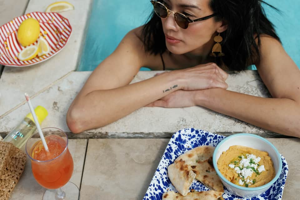 Woman in sunglasses enjoying tropical lunch poolside with hummus, pita bread, and fresh citrus drink