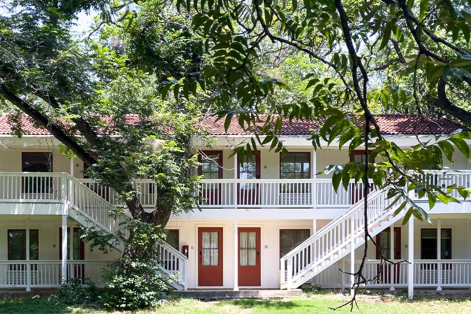 Two-story residential building with white railings and red doors under large trees