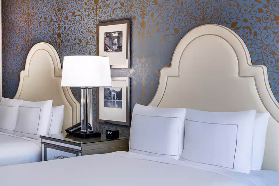 Luxury hotel bedroom with two queen beds, decorative blue and gold damask wallpaper, cream headboards, and framed artwork.
