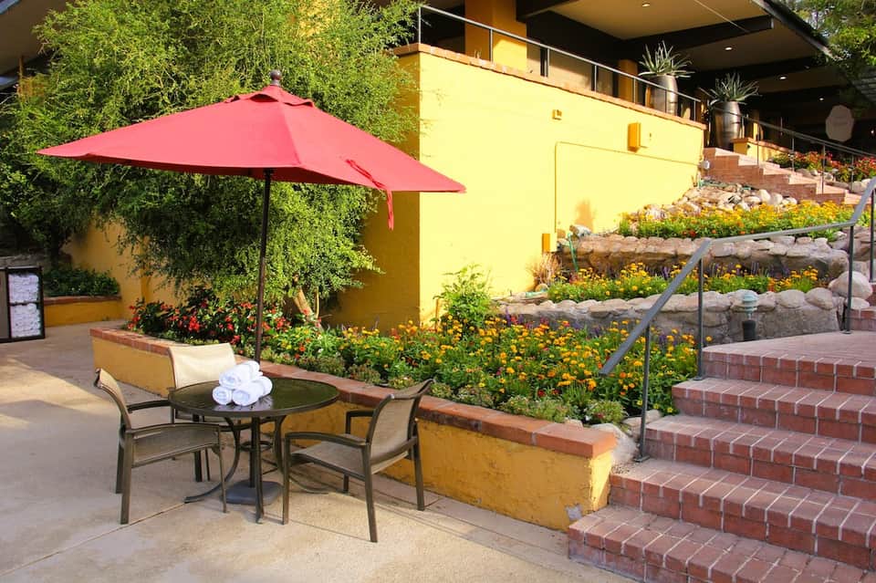 Outdoor patio with red umbrella, dining table, yellow building, and flowering gardens