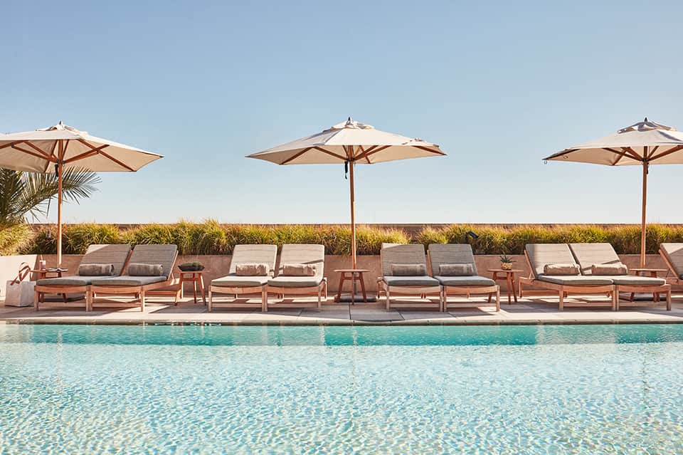 Resort pool area with tan loungers, beige umbrellas, and coastal grassland landscape under clear blue sky