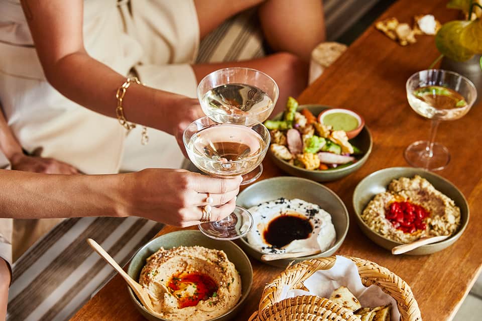 Two people toasting with cocktails at a wooden table with hummus bowls, salads, and appetizers