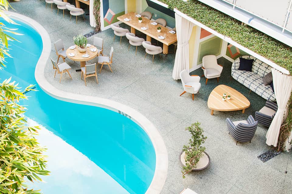 Aerial view of resort pool area with dining tables, lounge seating, cabanas, and surrounding landscaping.