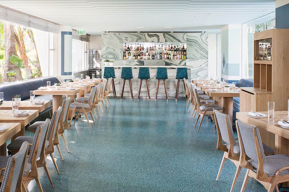Modern hotel dining room with teal bar stools, wooden tables, marble bar backdrop, and floor-to-ceiling windows