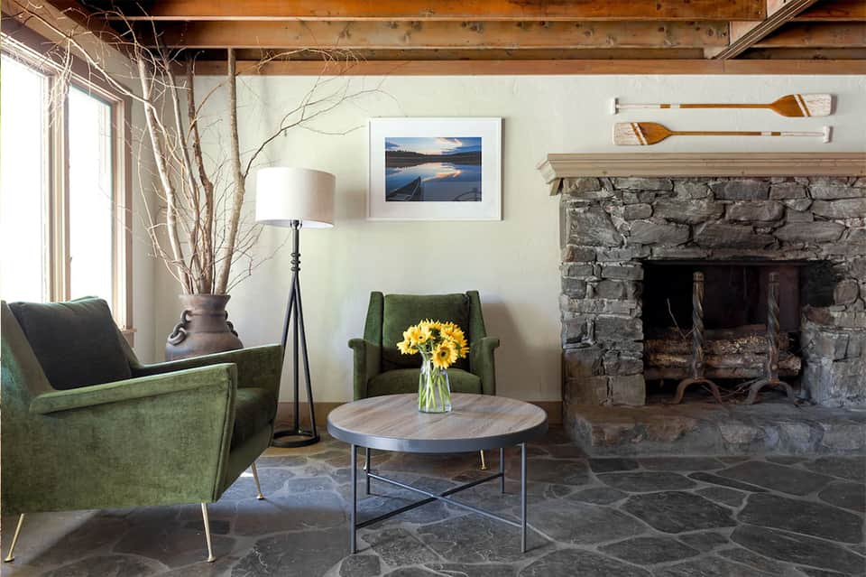 Rustic lounge with green chairs, fireplace, and stone hearth facing large windows with natural branches in vase
