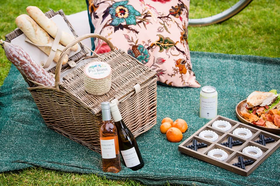 Outdoor picnic setup on grass with wicker basket, wine bottles, bread, fruit, and gourmet food