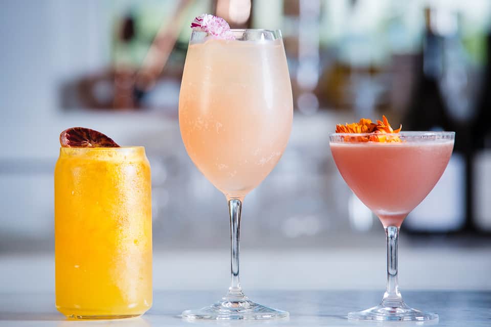 Three colorful cocktails on white surface: yellow drink, pink drink with flower garnish, and coral drink
