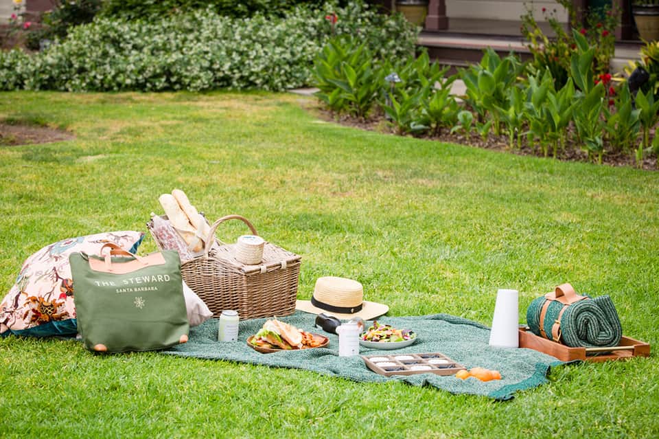 Picnic setup on grass with wicker basket, hat, food, and green blanket near garden landscaping