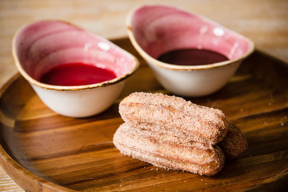 Cinnamon sugar-coated pastries with pink bowls of red dipping sauce on wooden board